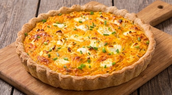 Una tarta salada redonda con relleno anaranjado y trozos blancos de queso, decorada con cebollino picado, sobre una tabla de madera en una mesa de madera.