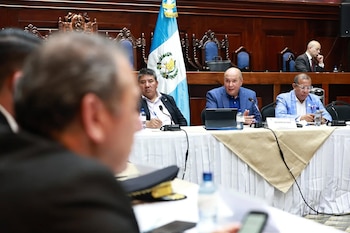 Vista de una reunión oficial con varios hombres sentados alrededor de una mesa, con una bandera de Guatemala prominente al fondo. Un cartel identifica a Orlando Blanco