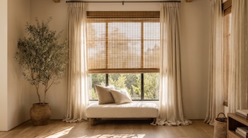 Sala con ventanal, persianas de madera, cortinas claras, chaise lounge con cojines, planta en maceta grande, piso de madera, muebles de madera, canasta tejida.