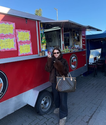 Una mujer con abrigo de piel marrón, gafas de sol y un bolso de mano posa frente a un food truck rojo. Se ven carteles con menú y paquetes de aperitivos