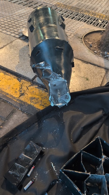 Cilindro de gas oscuro, abollado y roto en un extremo, sobre superficie negra en acera con líneas amarillas. Baterías y escombros plásticos cercanos