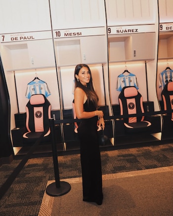 Antonela Roccuzzo, mujer de pelo oscuro y vestido largo negro, sonríe en un vestuario. Detrás, taquillas con los nombres de Messi y Suárez y camisetas de fútbol