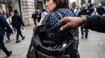 Una mano saca un teléfono celular negro del bolso abierto de una mujer que camina por la calle entre otras personas.