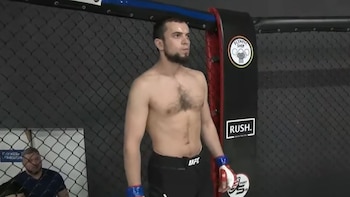Un hombre con barba y sin camiseta permanece de pie dentro de una jaula de combate con guantes de protección