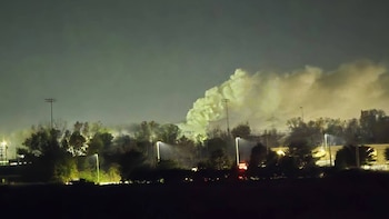 Una densa columna de humo, árboles, postes con focos de luz encendidos y un cielo oscuro nocturno