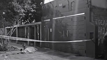 Fotografía en blanco y negro de una escena nocturna. Una calle con cinta policial que delimita el área. Un cuerpo cubierto en el suelo. Fondo de casas y árboles