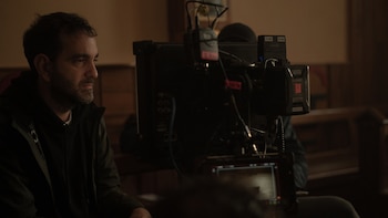 Nicolás Gil Lavedra, con barba, de perfil, observa una cámara de video profesional. Viste una chaqueta oscura. El fondo es un interior oscuro con madera