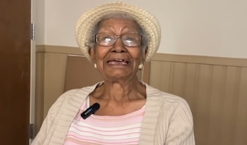 Retrato de Susan Young Browne, una mujer mayor con cabello gris, sombrero de paja, gafas y un micrófono prendido a su blusa a rayas