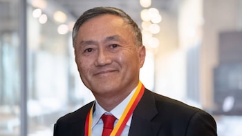 Primer plano de un hombre asiático de mediana edad sonriendo, con cabello gris, traje oscuro, camisa blanca, corbata roja y una cinta roja y amarilla alrededor del cuello