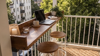Un balcón soleado con un escritorio de madera y taburetes. Hay un portátil plateado, una taza de café, velas y una planta en maceta.