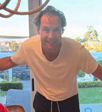 Un hombre de cabello castaño y camiseta blanca de cuello en uve sonríe delante de un ventanal con vista a un patio