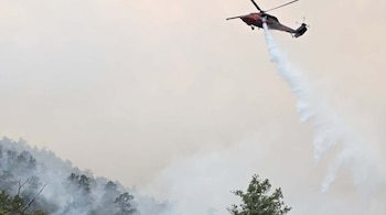Un helicóptero oscuro vuela sobre un bosque y descarga agua. El área está cubierta por una densa capa de humo entre los árboles
