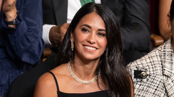 Primer plano de una mujer sonriente con vestido negro de tirantes y collar brillante, sentada en una sala junto a otras personas.