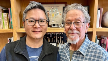 Primer plano de dos hombres sonriendo, uno asiático con gafas a la izquierda, y otro caucásico con barba y gafas a la derecha, frente a una estantería