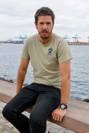 Un hombre con camiseta verde oliva y pantalones negros está sentado en un banco de madera junto al mar, con un puerto y grúas de carga al fondo