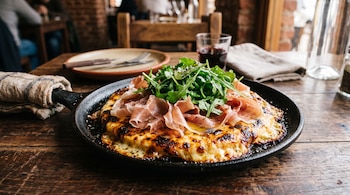 Sartén de hierro sobre una mesa de madera con queso provoleta derretido, jamón crudo, rúcula y un vaso con vino al fondo.