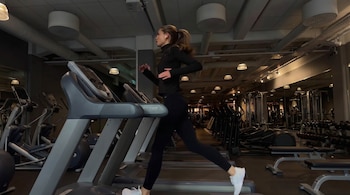 Una mujer con cola de caballo, ropa deportiva negra y auriculares blancos corre de perfil en una cinta de correr gris en un gimnasio bien iluminado.