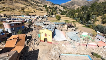 Vista aérea de un pueblo en una ladera de montaña con numerosos edificios derrumbados, escombros en las calles y personas transitando. Se observa una iglesia amarilla