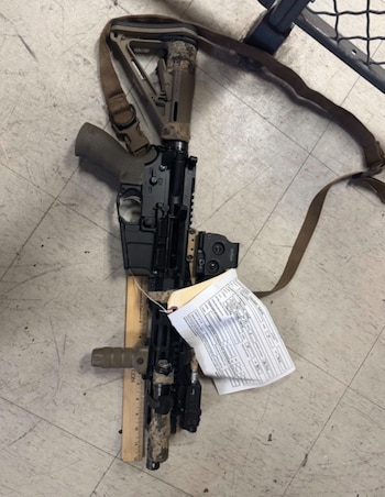 Un rifle oscuro con una mira, empuñadura vertical y una linterna sobre un suelo de baldosas grises. Un papel blanco con texto y una regla de madera están junto al arma