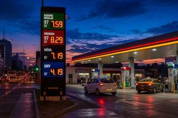 Un cartel luminoso muestra precios de combustibles junto a surtidores de una estación de servicio con dos vehículos estacionados al atardecer.