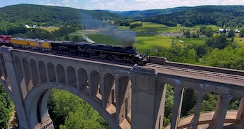 Una locomotora de vapor negra con la inscripción X4014 y una pequeña bandera estadounidense avanza sobre vías en un entorno de árboles verdes
