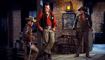 Tres hombres con sombreros de vaquero y atuendos western están de pie frente a una pared de ladrillos con un cartel y una lámpara encendida