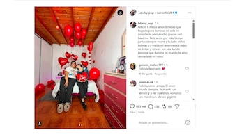 Dos mujeres en una cama. Habitación decorada con globos rojos redondos y de corazón. Una mujer sujeta almohadón Te Amo y besa mejilla de la otra