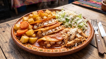 Plato de barro con tortillas dobladas en salsa roja, papas y zanahorias salteadas, lechuga picada, pollo deshebrado y cubiertos al lado.