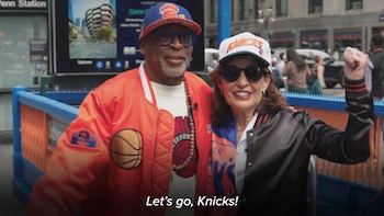 Hombre con gorra azul y naranja de los Knicks y chaqueta naranja junto a mujer con gorra blanca de los Knicks y chaqueta negra en la entrada de un metro