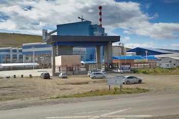 Edificios industriales azules y una chimenea a rayas rojas y blancas en Yacimientos Carboníferos Río Turbio, con aparcamiento de tierra y vehículos