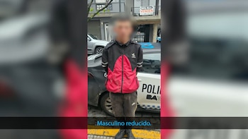 Un hombre de pie con el rostro difuminado y las manos detrás de la espalda frente a un vehículo policial en una calle urbana