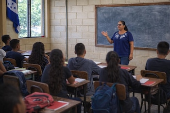 Maestra de pie en aula de Honduras, gesticulando frente a pizarrón vacío. Estudiantes sentados de espaldas en pupitres de madera. Bandera hondureña visible.