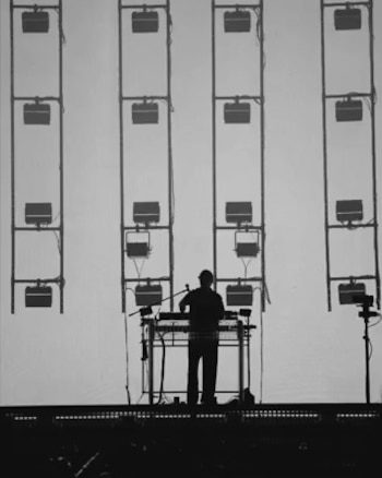 Silueta de un músico de pie en un escenario frente a una consola. Detrás, cuatro estructuras verticales con múltiples luces cuadradas