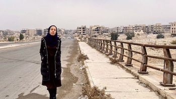 Una mujer con un hiyab azul y un abrigo oscuro camina por una carretera desierta junto a un puente oxidado, con edificios destruidos al fondo