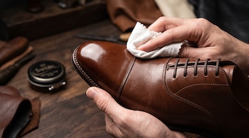 Manos sostienen un zapato de cuero marrón y limpian la puntera brillante con un paño blanco sobre una mesa de madera.