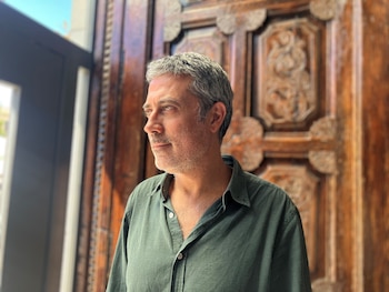 Primer plano de Andrea Bajani, un hombre de mediana edad con cabello y barba grises, camisa verde oscura, mirando hacia la izquierda. Al fondo, una puerta de madera tallada
