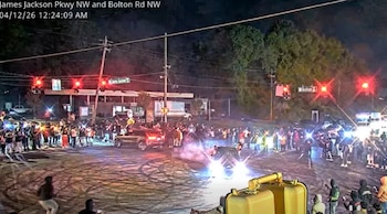 Imagen nocturna de una concurrida intersección en Atlanta donde vehículos realizan acrobacias con una multitud observando; se aprecian luces y chispas