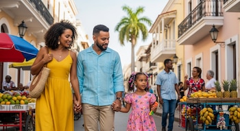 Una familia de tres personas, padre, madre e hija pequeña, camina por una calle empedrada con edificios de colores y puestos de frutas tropicales en un día soleado.