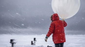 Persona vestida con parka roja sostiene un globo blanco grande en un campo cubierto de nieve bajo la tormenta.