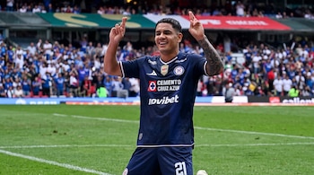 Un futbolista con uniforme azul marino del club Cruz Azul está de rodillas en la cancha haciendo un gesto con los dedos cruzados.