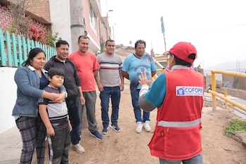 Un representante de COFOPRI con chaleco rojo y gorra roja habla a un grupo de seis residentes (cuatro hombres, una mujer y un niño) en un área urbana