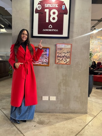 Carolina Baldini, de pie, sonríe y señala con su mano derecha una camiseta de fútbol enmarcada de color granate con el nombre Simeone y el número 18