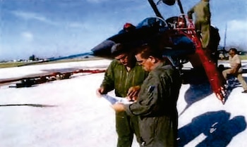 Dos hombres en uniformes de vuelo o trabajo, Luis Raúl González-Pardo Rodríguez y Lorenzo Alberto Pérez-Pérez, examinan documentos en una pista con un helicóptero detrás