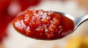 Cuchara de metal con kétchup rojo que contiene cristales de azúcar transparentes y pequeñas partículas de tomate. Fondo borroso en tonos rojo y blanco.