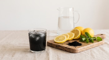 Vaso de limonada negra con hielo, limones cortados y enteros, carbón activado en polvo, hojas de menta, tabla de madera, jarra de agua y mantel beige.