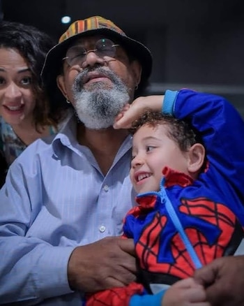Un hombre mayor con sombrero, gafas y barba canosa abraza a un niño con chaqueta de Spider-Man, junto a una mujer que sonríe al fondo