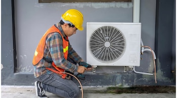 Hombre arrodillado con casco amarillo y chaleco naranja instala una unidad de aire acondicionado blanca en una pared gris, usando un taladro eléctrico.