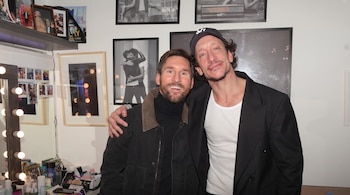Lionel Messi con pulóver negro y Nico Vázquez con gorra posan sonriendo. Al fondo se ven espejos con luces y cuadros en una pared.