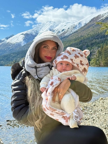 Una mujer con abrigo gris con capucha sostiene en brazos a un bebé abrigado frente a un lago y montañas con nieve