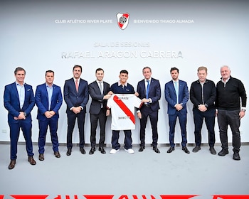 Diez hombres de pie, uno sostiene una camiseta de fútbol blanca con franja roja y nombre Almada. Fondo blanco con escudo de River Plate y texto